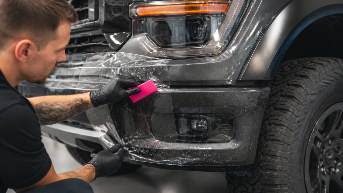 Close-up of a technician in a clean shop applying a clear paint protection film panel to the front bumper of a pickup truck, squeegee in hand, water droplets visible on the film